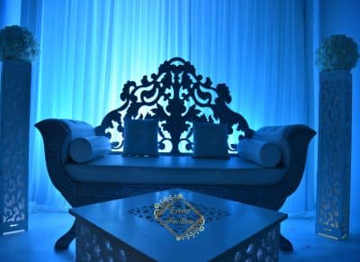 Scène de mariage luxueuse éclairée en bleu électrique. Banquette baroque ornée, table basse sculptée et socles floraux devant un rideau lumineux.