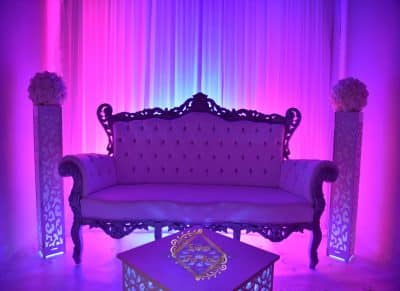 Canapé baroque blanc pour un lounge VIP, flanqué de piliers lumineux sculptés et de fleurs, sous un fond de rideaux violets et magenta.