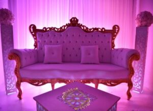 Luxueux canapé baroque rose en velours capitonné, éclairé en magenta. Décor de scène, deux colonnes florales et table décorée.