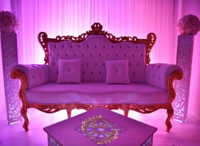 Luxueux canapé baroque rose en velours capitonné, éclairé en magenta. Décor de scène, deux colonnes florales et table décorée.