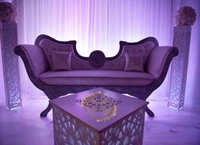 Salon oriental violet événement. Canapé baroque en velours, table lumineuse à motifs géométriques et piliers fleuris devant des rideaux drapés.