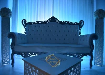 Élégant canapé des mariés blanc ornementé, ambiance événementielle bleu profond. Décoration avec colonnes fleuries et table basse logo doré.