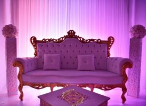 Causeuse royale baroque violette et or capitonnée. Décor de mariage élégant avec éclairage magenta sur rideaux et deux colonnes florales.