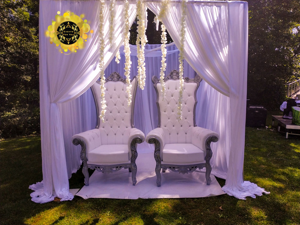 Luxueuse décoration de mariage extérieur. Deux chaises trônes blanches capitonnées sous un drapé blanc fleuri. Logo Events For You.