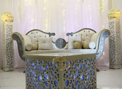 Dias mariage (Encore Un Rêve). Canapé ivoire capitonné argenté, table géométrique lumineuse bleue. Poteaux décoratifs à motifs marocains.