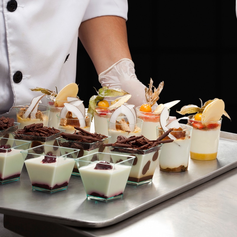 Verrines de dégustation : 15 desserts miniatures gourmands Plateau de verrines de dégustation : panna cotta, mousses et desserts chocolatés, décorés de physalis et noix de coco. Chef en arrière-plan.