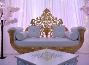 Sofa trône luxueux doré et blanc avec dossier sculpté, devant des rideaux lumineux roses. Table basse décorée.