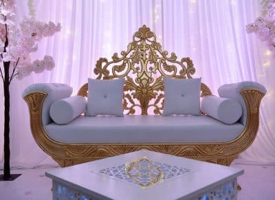 Sofa trône luxueux doré et blanc avec dossier sculpté, devant des rideaux lumineux roses. Table basse décorée.