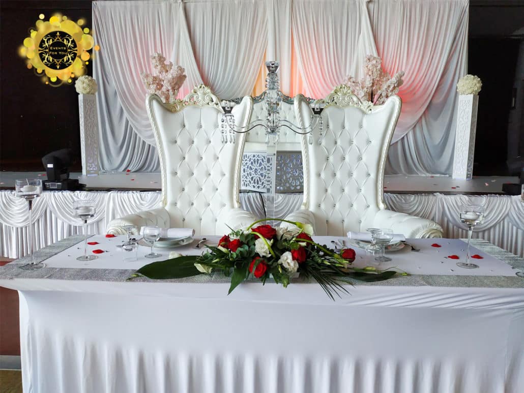 Table d'honneur de mariage luxueuse avec deux chaises trône blanches capitonnées, décor floral rouge et blanc, et rideaux élégants.