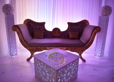 Trône de mariés baroque pourpre et or. Décoration de mariage luxueuse avec table basse marocaine éclairée et colonnes fleuries sous un éclairage violet.