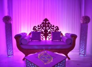 Scène de mariage romantique: trône royal pourpre, colonnes lumineuses et tentures blanches. Table basse affichant "Excellence For Your Day".